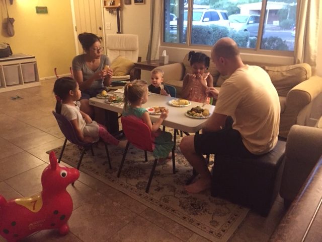 Two adults sit with four children at a small table and eat a meal.