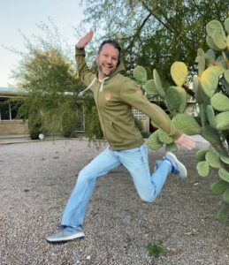 Corey Gilchrist in a green sweater in front of a green cactus jumping with joy.